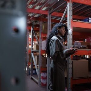 An adult woman in uniform checking inventory in a warehouse aisle.