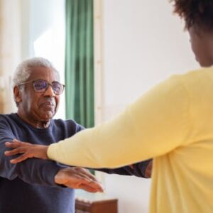 An elderly man with a caregiver assisting him in exercise at home.
